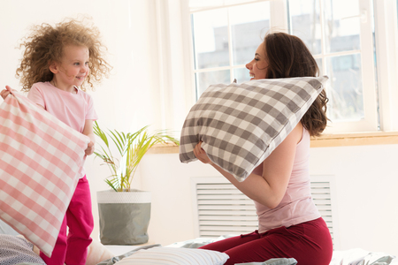 Family morning. Daughter with curly-haired mother and divide pillows on bed in bedroom. They fool around and arrange pillow fight. Happy childhoodの写真素材