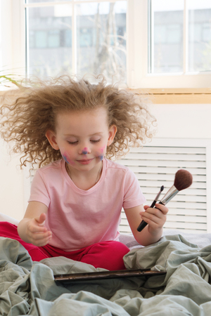 Little girl and mother's make-up. She is holding makeup brush, palette with shadows and mirror. Child's face in paint. Positive emotions and happy childhood.の写真素材