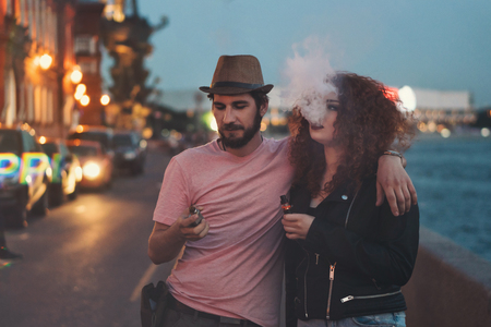 Loving couple of hipsters on date. Guy in hat and T-shirt is holding electronic cigarette and hugging girlfriend. Girl with curly red hair smokes an ecigarette. They are in streets of evening city.