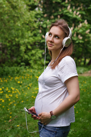 Young pregnant girl listening to music on headphones. She is holding a cell phone in her T-shirt and jeans. Online musicの写真素材