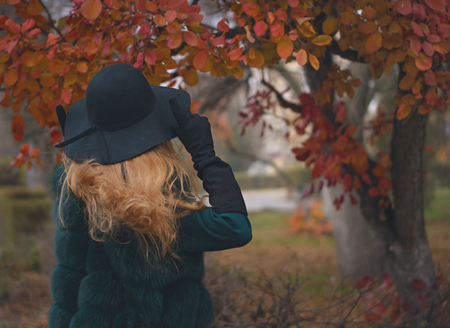 Red-haired girl in autumn park. She's wearing short coat, wide-brimmed hat and gloves. Portrait from back. Man is unrecognizable.の写真素材