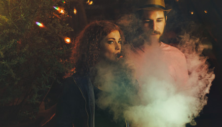 Loving couple on date. Guy and girlfriend smoke electronic cigarettes. Clouds of smoke. Red-haired...