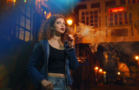 Young attractive red-haired girl smokes electronic cigarette. She walks through small streets of city. Evening city lights glare.の写真素材