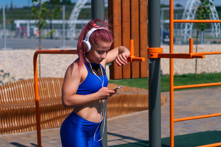 Girl on street workout. Young slender woman listening to music on headphones. She is holding cell...
