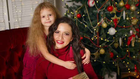 Family celebration of Christmas. Daughter hugs her mother tightly by shoulders. In background is Christmas tree. Happy childhood.の写真素材