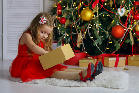 Little girl in red dress sits on carpet and opens a gift. Christmas tree with ornaments in the background. Happy childhood.の写真素材