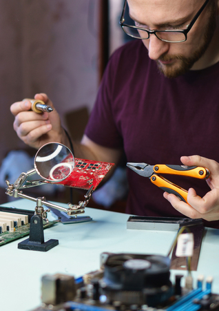 Repair motherboard. Man at work. He is holding a soldering iron and pliers. Electronic card under...