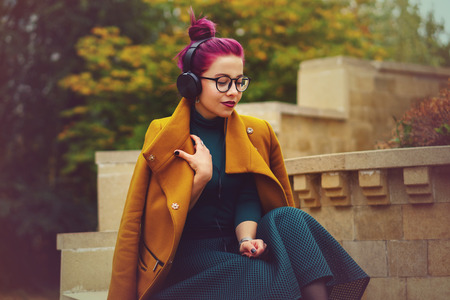 Cute young girl listening to music on headphones in autumn park. Girl has eggplant hair. Woman enjoys melody. She is wearing glasses and mustard-colored coat.の写真素材