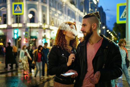 Couple in love on date. Boyfriend holds phone. She hugs him and smokes an electronic cigarette. They walk streets of night city. Wet asphalt after rain.の写真素材