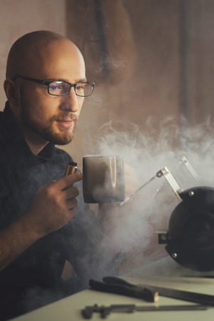 Master to break smoking and drinking hot beverage. Man in glasses in thought holds mug in his hands and lets out smoke.の写真素材