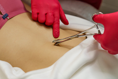 Beautician makes puncture to navel of thin girl in spa salon. Hands of master hold special tongs.の写真素材