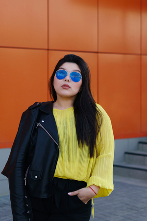 Portrait of a young brunette plus size woman. Audacity pan-Asian girl in a biker leather jacket with sunglasses poses near a bright wall. Body positivity movement. Street fashion. Leisure timeの写真素材