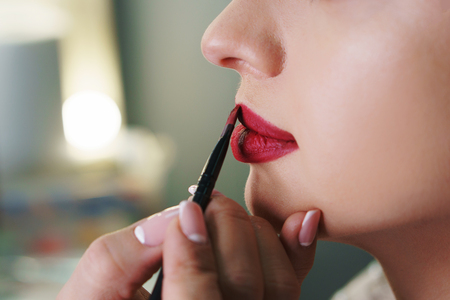 Makeup artist working in makeup studio, applies red lipstick to lips with brush. Female face close up side view.の写真素材