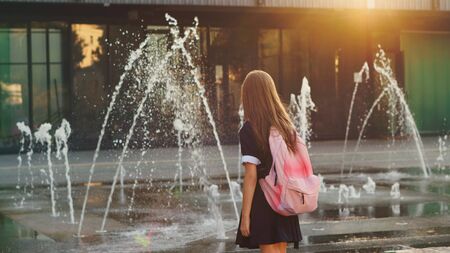 Young schoolgirl with backpack on her shoulders on way home. She stopped to admire fountain. Full-back position.の写真素材