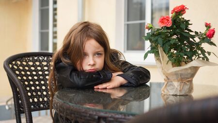 Fashionable little schoolgirl sitting at table of summer terrace of restaurant. She's upset about...