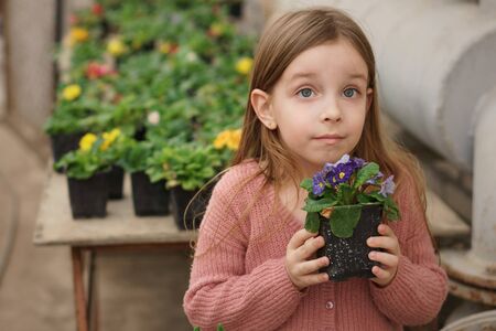 Cheerful schoolchild holding pot with blooming plant in hands in orangery. Excursion in greenhouse. Summer girl fashion. Happy childhood. Natural beauty. Childrens day. Botanical. Blooming gardenの写真素材