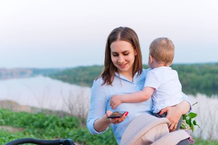 Young loving mother walking with her little son on open air. Mompreneur checks email. Career girl combines child care and work. Family time on nature. Hiking and adventure with little kid.の写真素材