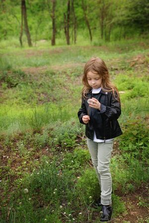 Stylishly dressed blonde little girl schoolgirl is trying to save a white dandelion. She stands in a green glade. Walk in the park.の写真素材