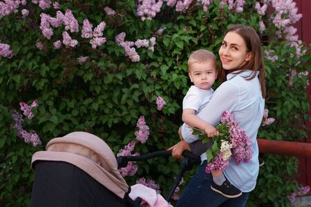 Young caring mother on walk with two children on background of spring flowering. Eldest child in her arms is sitting in her mothers arms. Baby is sleeping in stroller. Happiness to be parent.の写真素材
