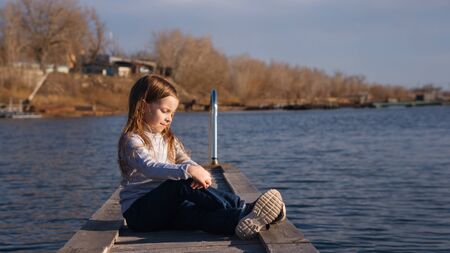 In harmony with nature, alone with himself. A little girl sits on a wooden pier near the lake. The sun blinds the eyes. Campaigns and adventures. Happy childhood, lifestyle concept.の写真素材