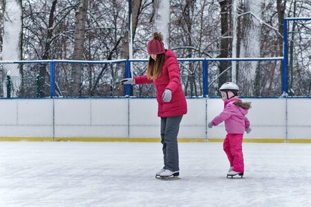 Little adorable girl with her mom learning to skate. Family skating and training with white skates on the ice area in winter day. Weekends activities outdoor in cold weather.の写真素材