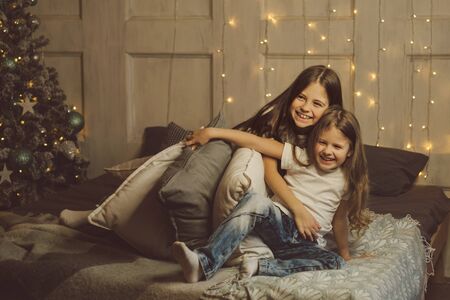 Little friendly sisters fight pillows. Morning light Christmas holiday. Children play and have fun together waiting for presents for the holiday. Room in bright christmas lights. Holiday decorations.の写真素材