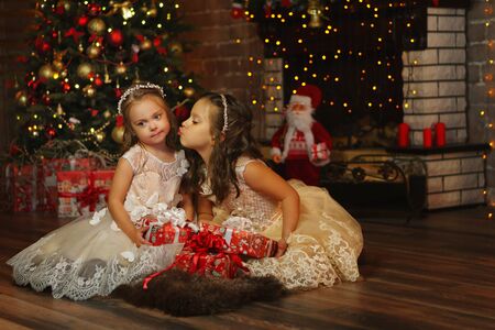 Little charming sisters in luxurious elegant dresses exchange gifts on Christmas Eve. Room in bright christmas lights. Holiday decorations. Related feelings. Friendly family. Spending time togetherの写真素材
