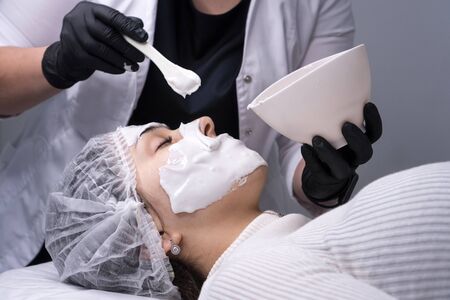Cosmetologist applies alginate mask with spatula on face of woman. Facial skin treatment in beauty...
