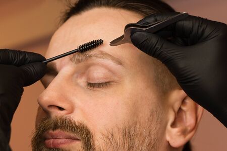 Professional barber doing threading procedure and correcting shape of eyebrows to young male client with tweezer in barber shop. Barber at work. Brow care concept. Plucking browsの写真素材