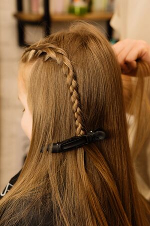 Process of braiding the master weaves braids on her head blond little girl in beauty salon close up....