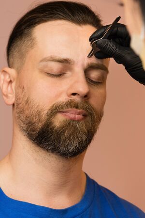 Professional barber doing threading procedure and correcting shape of eyebrows to young male client with tweezer in barber shop. Barber at work. Brow care concept. Plucking browsの写真素材