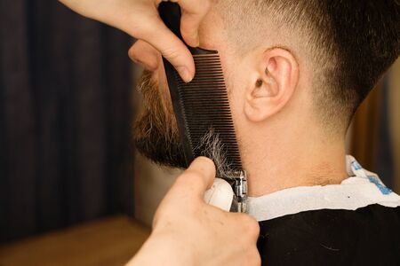 Barber trimming beard of male customer with electric razor at barbershop. Man with beard and moustache getting stylish trendy hair shaving, with hair clipper and comb. Close up.の写真素材