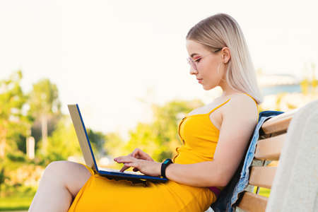 Young attractive girl in yellow dress works at laptop while sitting on bench in park. Concept of...