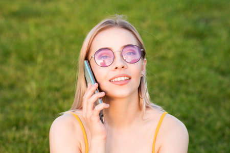 Close-up portrait of a girl in bright sunglasses talking on phone on background of green grass on a...