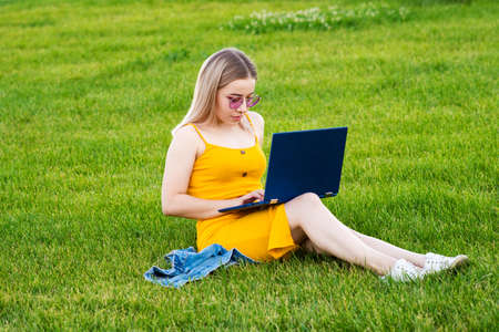 Woman is using a laptop in a green field. Freelance Worker Concept. Girl with laptop outdoors. A...