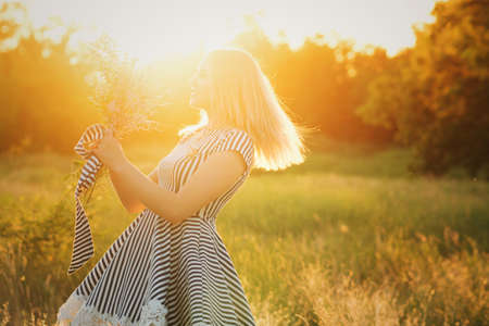 Portrait eco-friendly woman in striped sundress on meadow with bouquet of wildflowers in hand dancing on green field in sunset and sun glare. Summer evenings in nature. Background. Space for textの写真素材