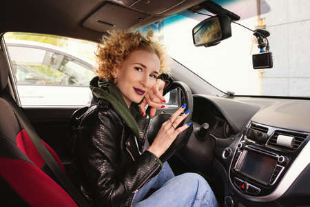 Portrait curly hair woman siting in her own automobile, keeping hands on wheel, wearing a rock black style. People, driving, transport concept. Sideways shot. Urban fashion conceptの写真素材