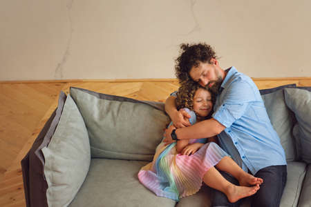 Portrait Young Friendly Family. Father and daughter relaxing in sofa. Family Time Together. Daddy Hugs Daughter with Tendernessの写真素材