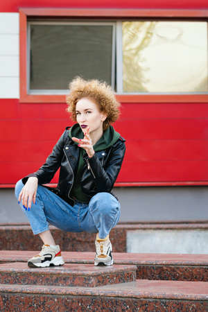 Full length portrait of trendy hipster girl sitting at the wall background. Urban fashion concept....