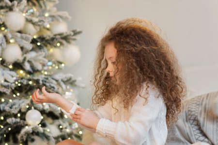 Christmas morning, cute little girl with curly hair looking at the gifts in the background of the Christmas tree. New year, christmas concept. Christmas eve at homeの写真素材