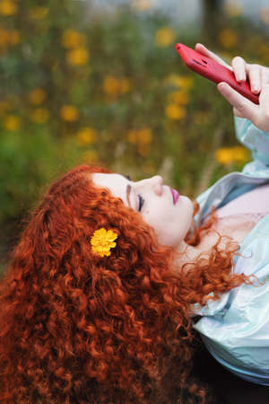 Romantic Curly Red-haired Girl Lying On A Bench Reading Internet Messages. Mobile Phone User. Online Internet Connection Concept.の写真素材