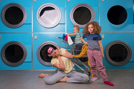 Mother Household With Children Wearing Pyjamas Having Fun And Doing Laundry At Self Service Laundrette Near Washing Machines. Housework Togetherの写真素材