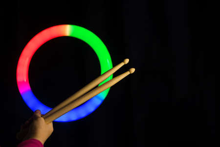 Child Hand Hold Wooden Drumsticks On Black Backlit RGB Circle Background. Rock Music Concept. Unrecognizable. Advertising Drum Lessons For Children. Soft Focusの写真素材