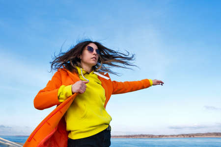 Atmospheric lifestyle photo Millennial Hipster Girl in trendy colorful casual outfit. Happy brunette young woman in good mood walks outside on pier next to sea.の写真素材