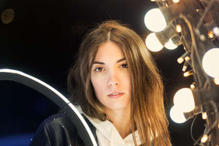 Millennial Hipster Girl Wearing Black Jacket Walking in City Street. Having Good Mood Over Glowing Festive Bokeh. Garlands Lights Urban Background.の写真素材