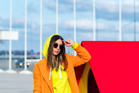 Millennial Hipster Girl in lilac sunglasses, orange coat, yellow hoodie posing at urban background....