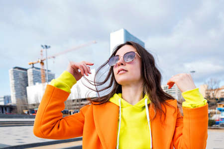 Happy brunette young woman in good mood walks outside and touches hair. Close up portrait Millennial Hipster Girl in lilac sunglasses, orange coat, yellow hoodie posing at urban backgroundの写真素材