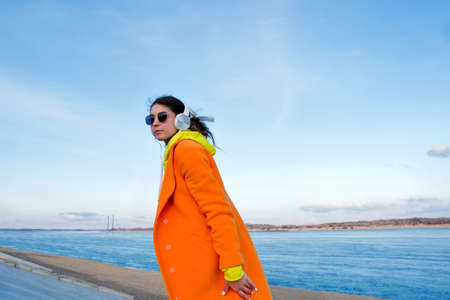 Good-looking young woman walks outside on pier next to sea. Real people. Hipster Girl listens to music in white headphones and looks at camera looking over his shoulderの写真素材
