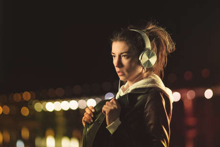 Millennial Girl Enjoying Nightlife In City Street. Listening Music With Headphones. Gorgeous Hipster Youngster Dancing In Colorful Neon Illumination Outdoors. Bokeh Urban Lightsの写真素材