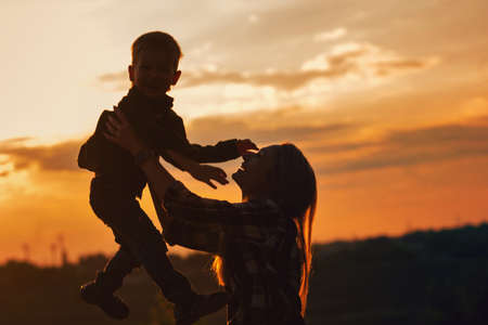 Silhouette young mother with pleasure spending time with kid in park. Lovely Mommy throw up son...